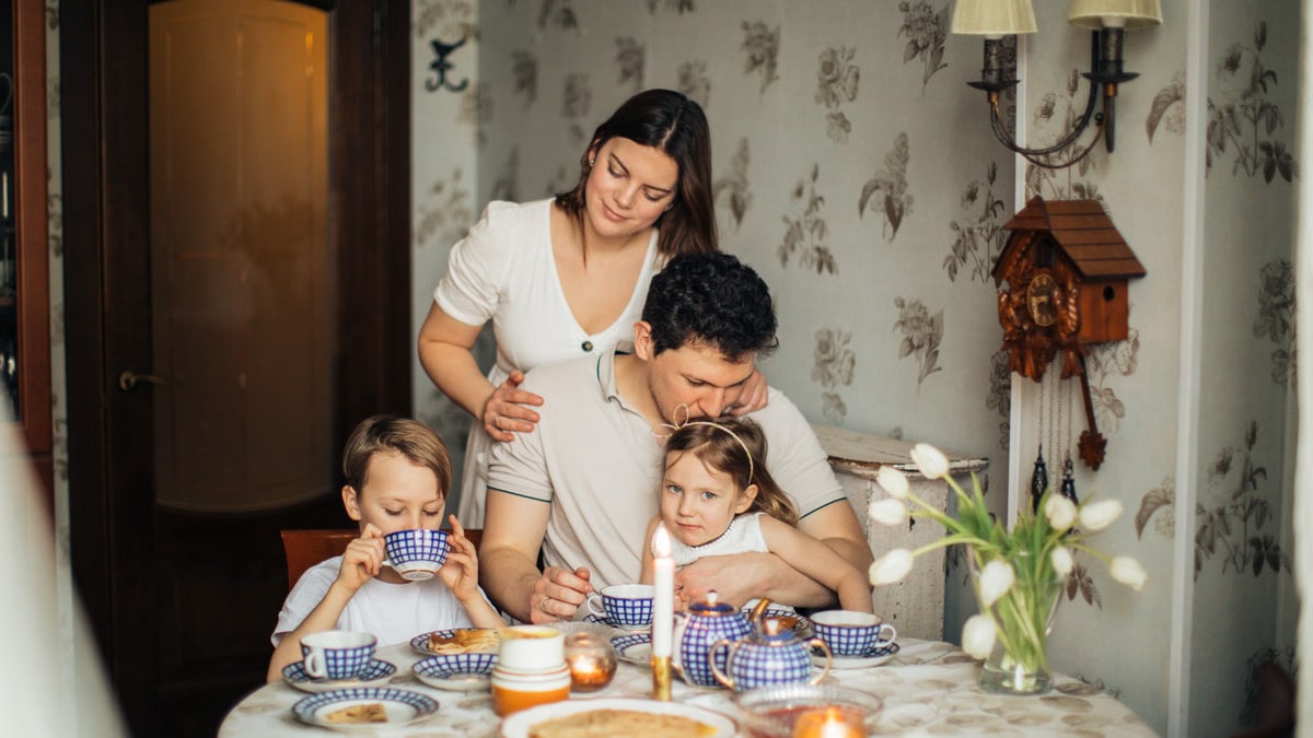 Happy family with children gathering at table with blue tea set having calm tea time with homemade pancakes in cozy living room